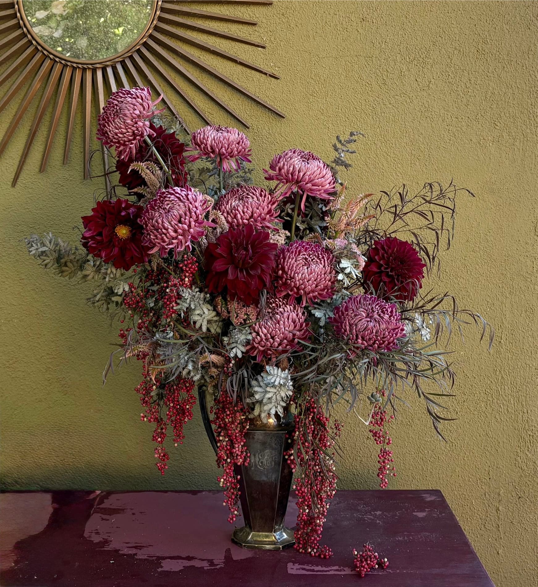 Burgundy chrysanthemums and pepperberry beneath a sunburst mirror on an olive wall